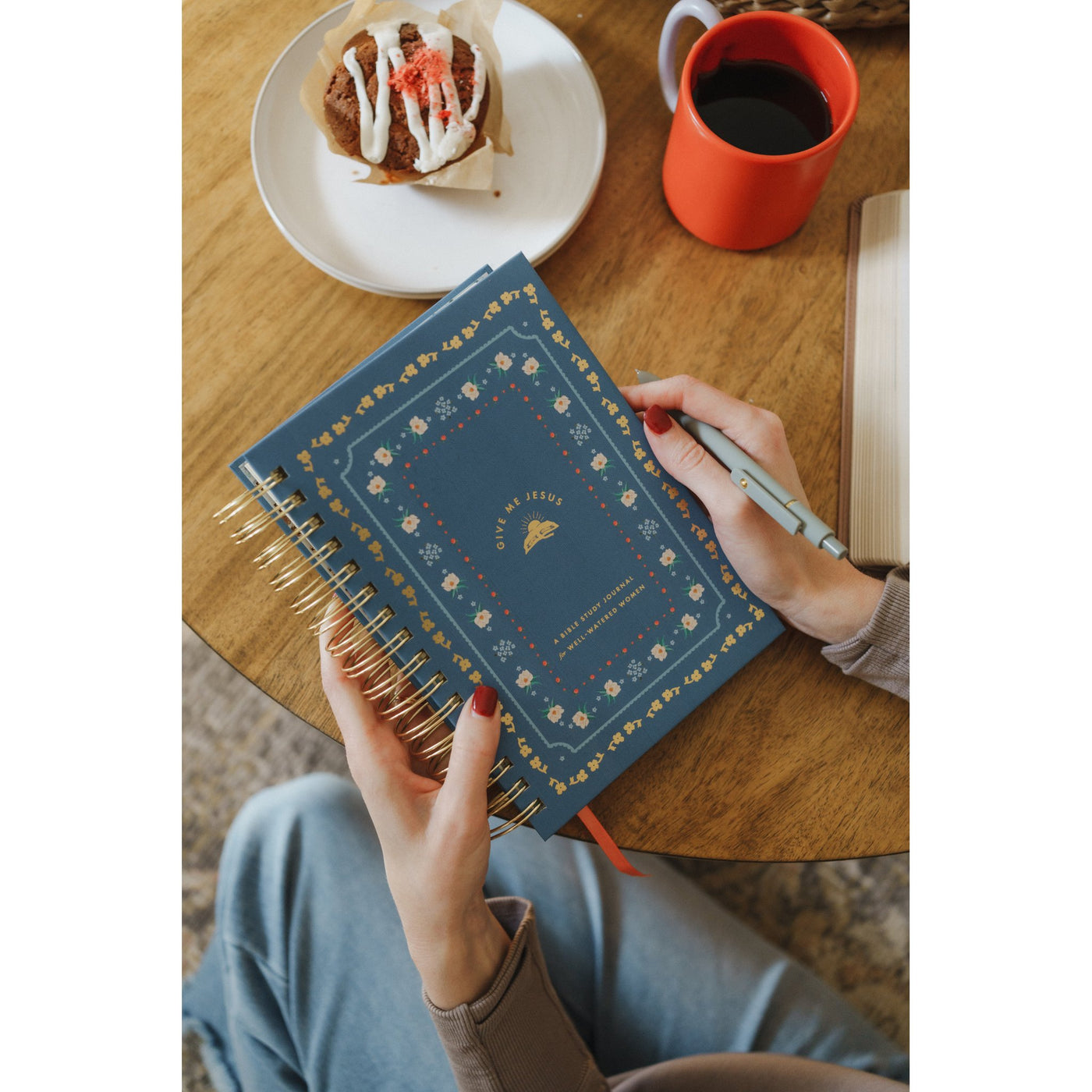 Hands holding the Give Me Jesus Journal next to coffee and a morning pastry on a wooden table.