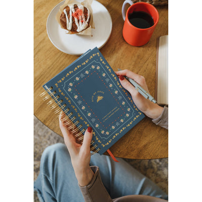 Hands holding the Give Me Jesus Journal next to coffee and a morning pastry on a wooden table.