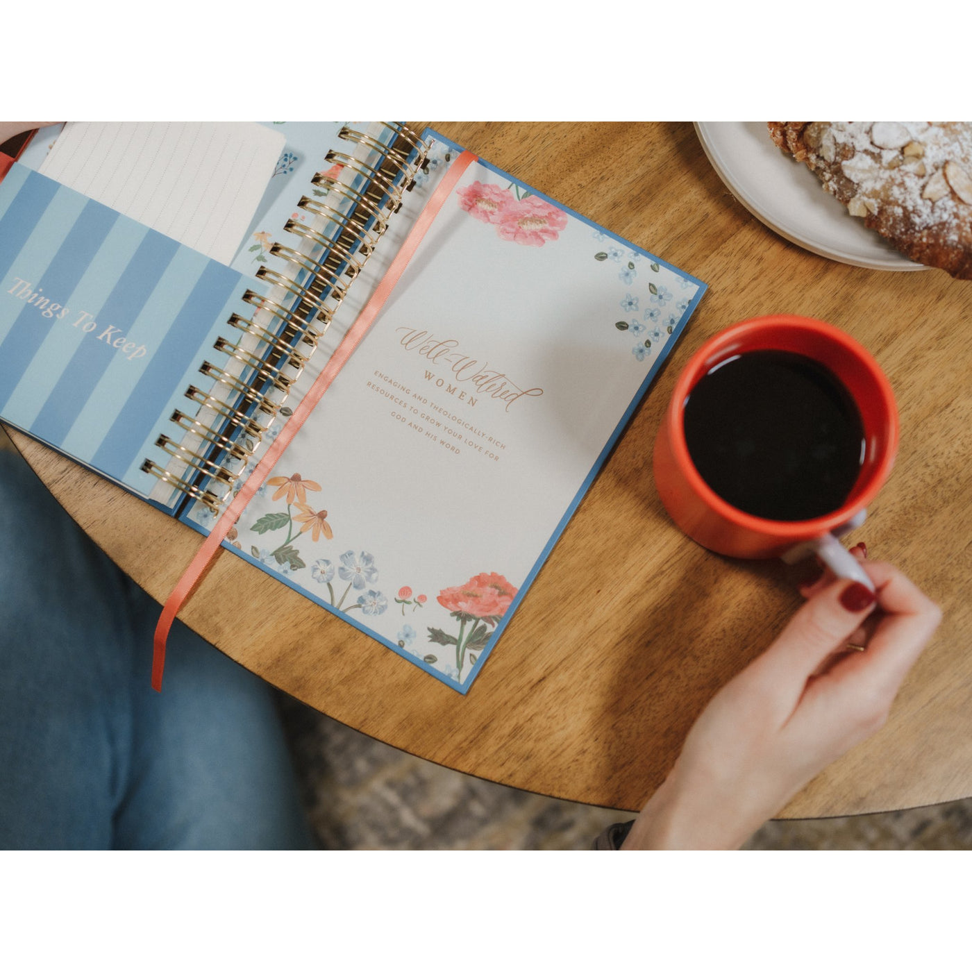 Hands holding the Give Me Jesus Journal next to coffee and a morning pastry on a wooden table.